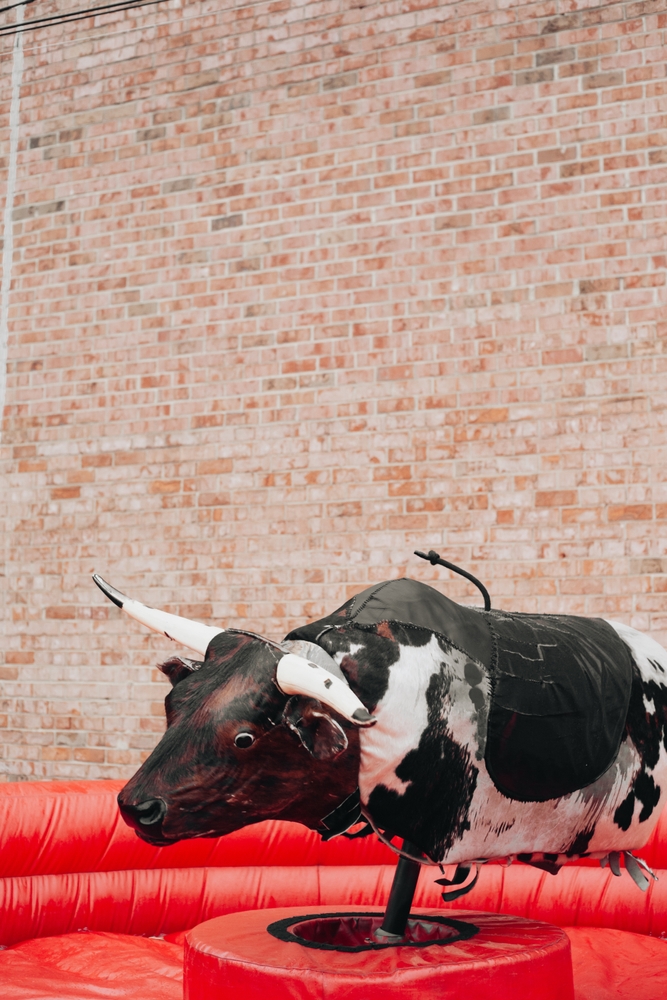 At a lively fair, a mechanical cow stands ready for bull riding. Enthusiastic participants take turns trying to stay on as it spins and bucks, surrounded by a vibrant red backdrop.